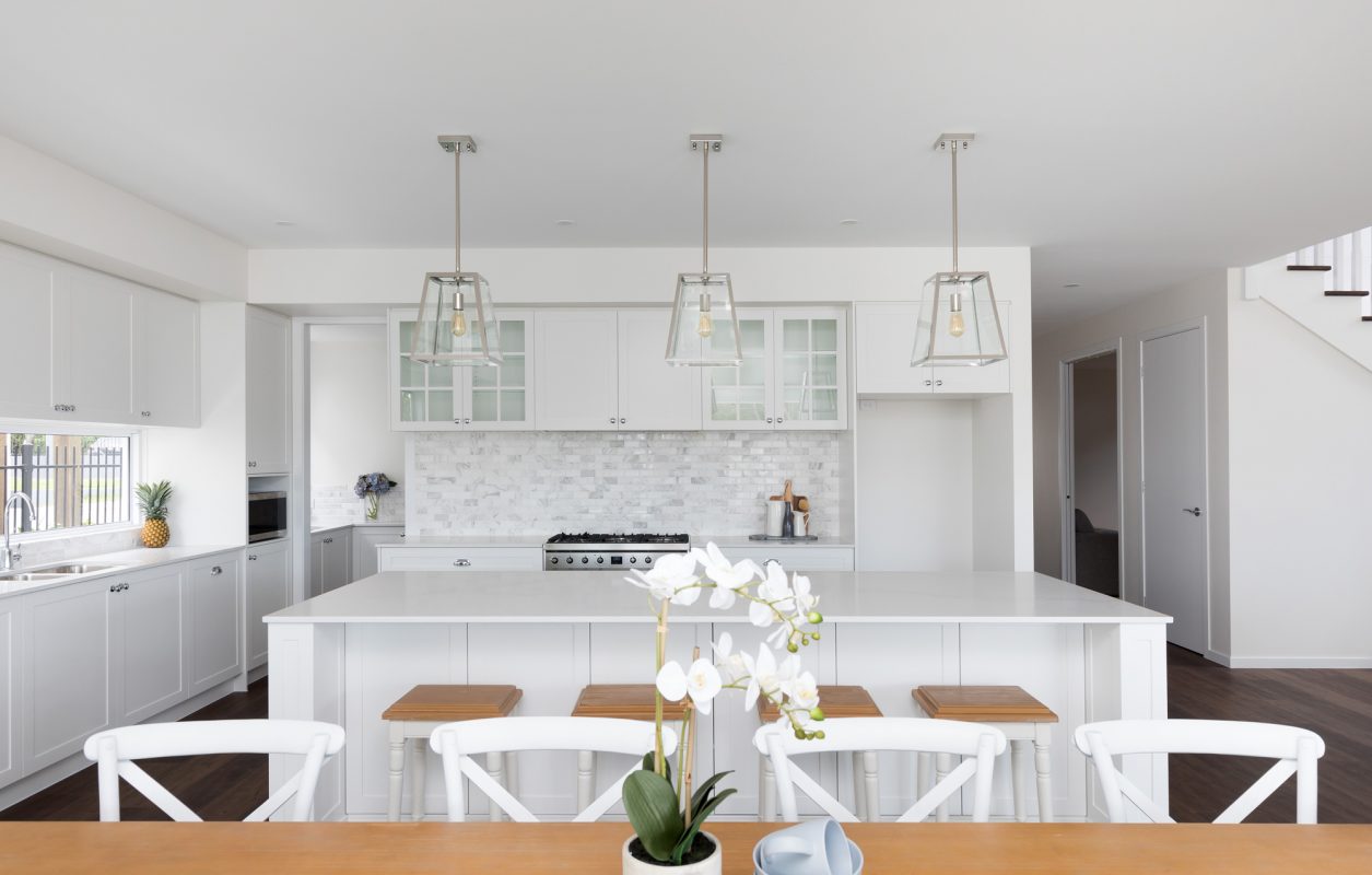 White open plan shaker kitchen with a light brown dining table in the foreground that has a vase with white orchids in it