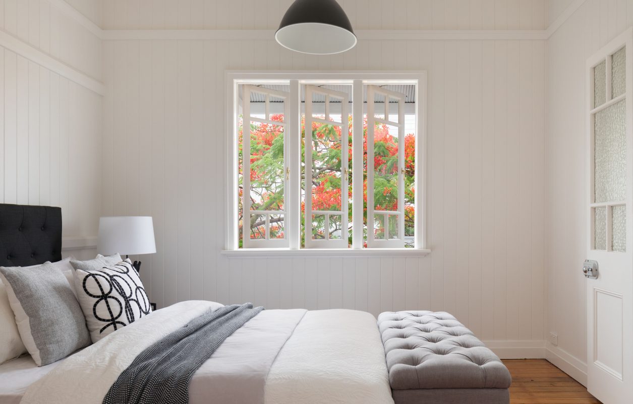 Cottage style bedroom with windows facing a flowering tree, white wooden walls, grey blanket box and a black headboard